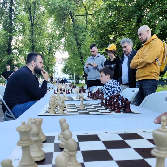 Šachmatų stalas iš arti. Tolumoje matosi susimąstę žaidėjai.