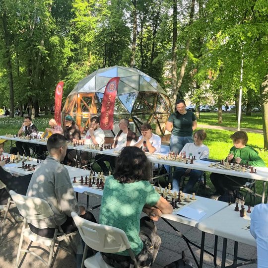 Dvi šachmatų stalų eilės, prie kurių sėdi žaidėjai.
