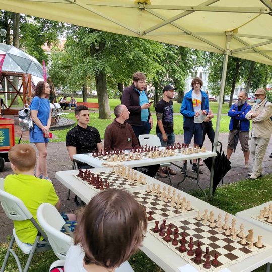 Po paviljonu prie stalų sėdi šachmatininkai. Minia žmonių stovi aplink juos.