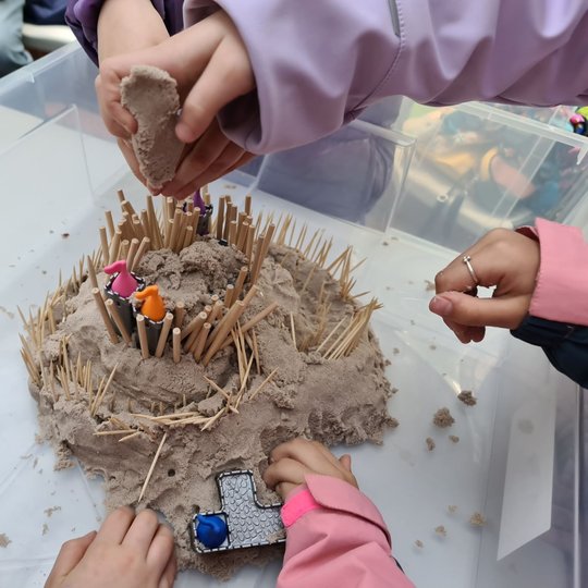 Vaikučiai rankomis stato smėlio pilį plastikinėje dėžėje.