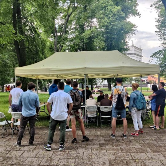 Po paviljonu prie stalų sėdi šachmatininkai. Minia žmonių stovi aplink juos.