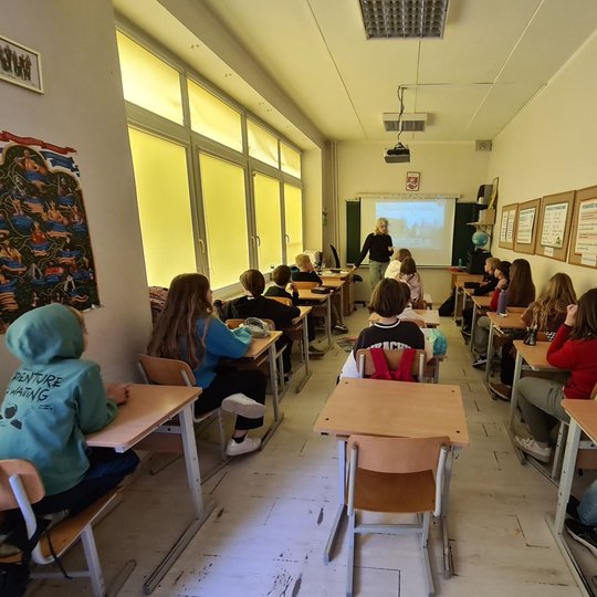 Maža ketvirtokų klasė, fotografuota iš galo. Klasės priekyje stovi mokytoja.