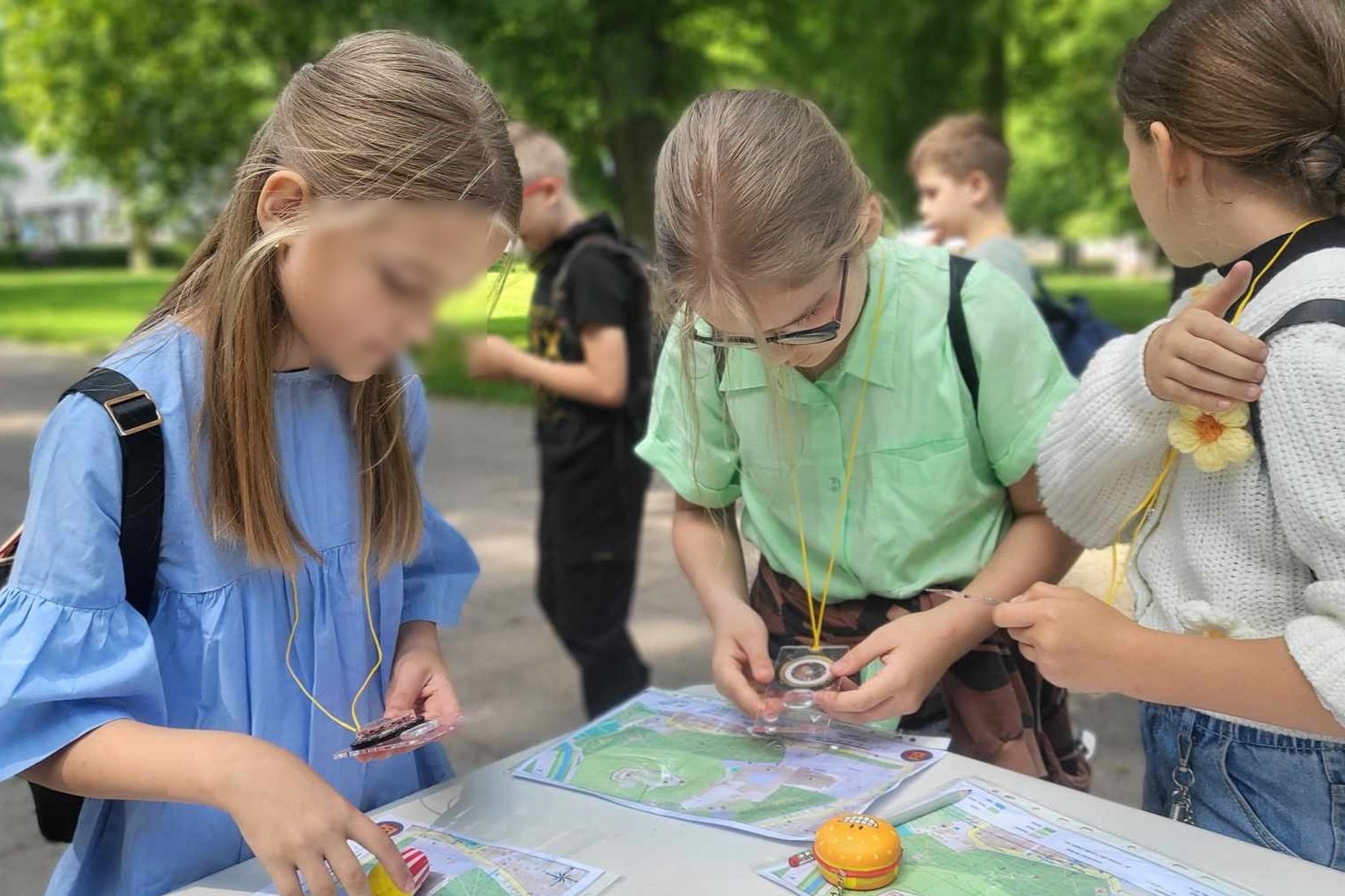 Trys mokinės stovi prie balto stalo parke, naudojasi ant kaklo esančiais kompasais ir tyrinėja popierinius pilių planus.