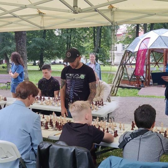 Vyksta šachmatų žaidimai, žmonėms susėdus prie stalų. Prie jų dar stovinėja keli žmonės.