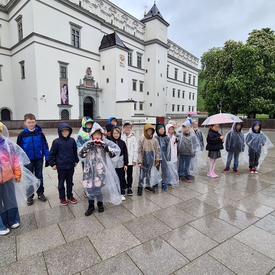 Vaikai su lietpalčiais pozuoja Katedros aikštėje, už jų Valdovų rūmai.