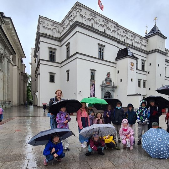 Mokiniai su mokytoja po skėčiais pozuoja prie Valdovų rūmų, Katedros aikštėje.