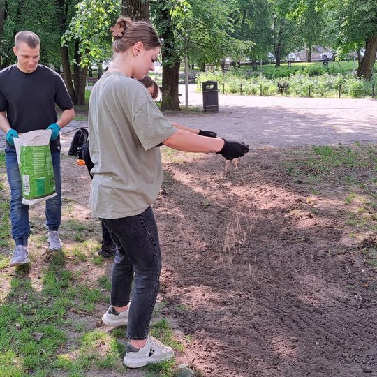 Vyksta sėjimo procesas - vyras neša sėklų maišą, moteris barsto sėklas ant supurento ir išlyginto tako.
