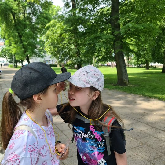 Dvi mokinės, primerkę po akį, žiūri viena į kitą per stiklinį kompasą.