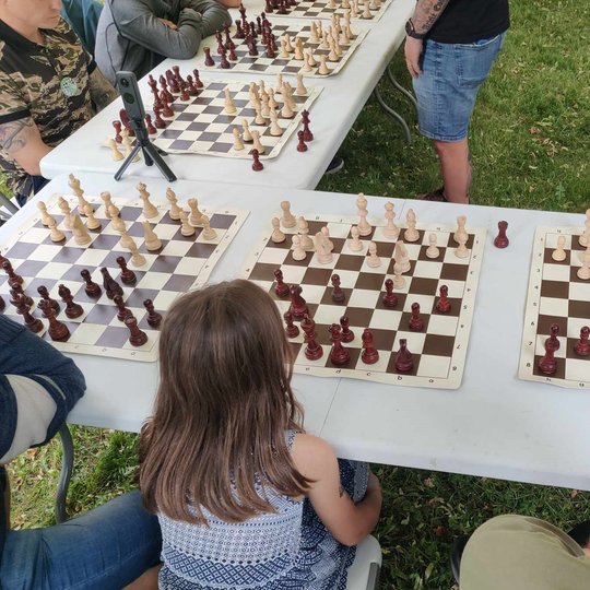 Šachmatų stalai, prie kurių nugara į objektyvą atsisukę sėdi žaidėjai.