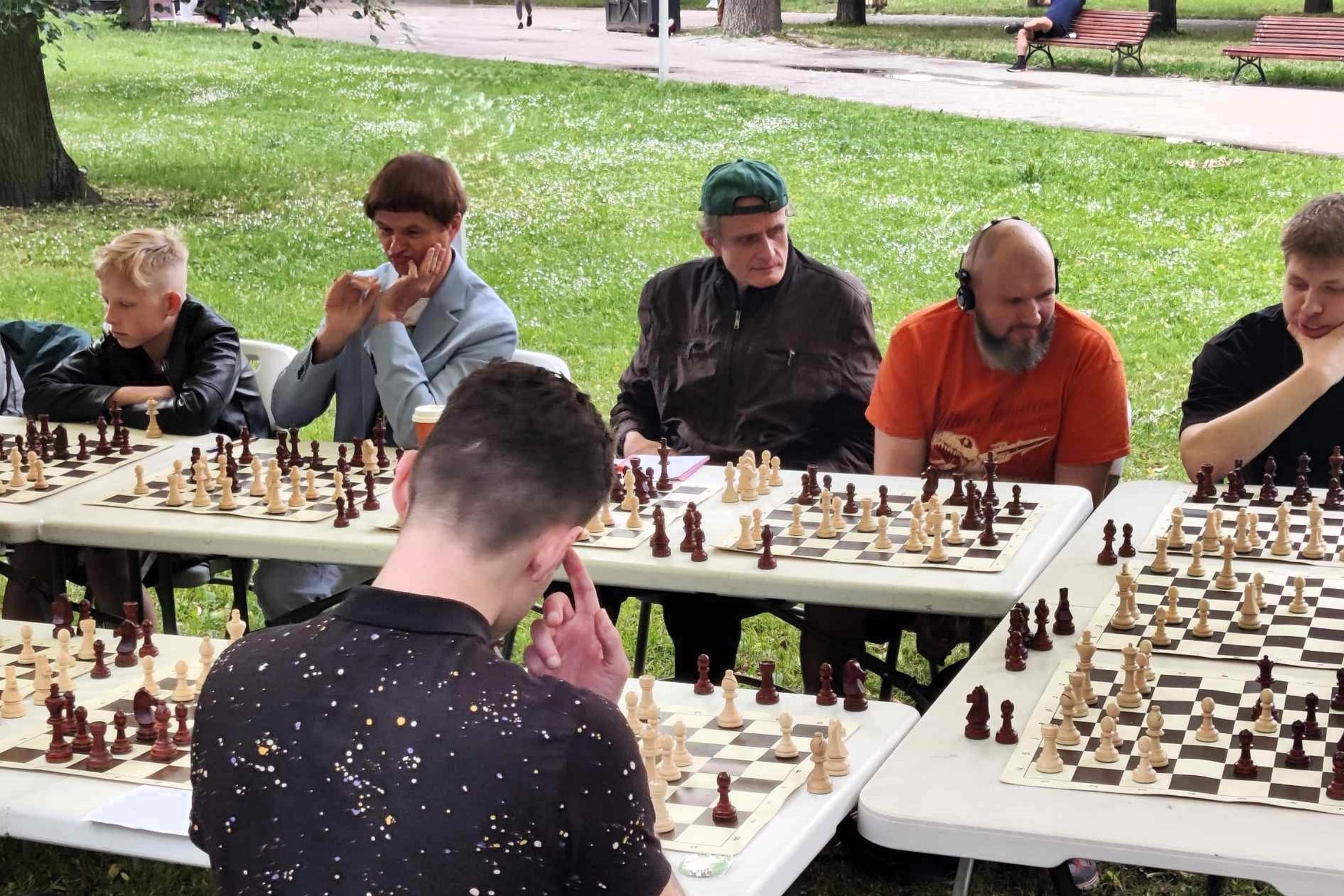Prie šachmatų stalų sėdi vyrai ir berniukas. Visi susimąstę.