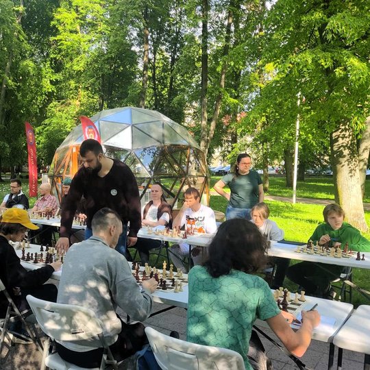 Dvi šachmatų stalų eilės, prie kurių sėdi žaidėjai.
