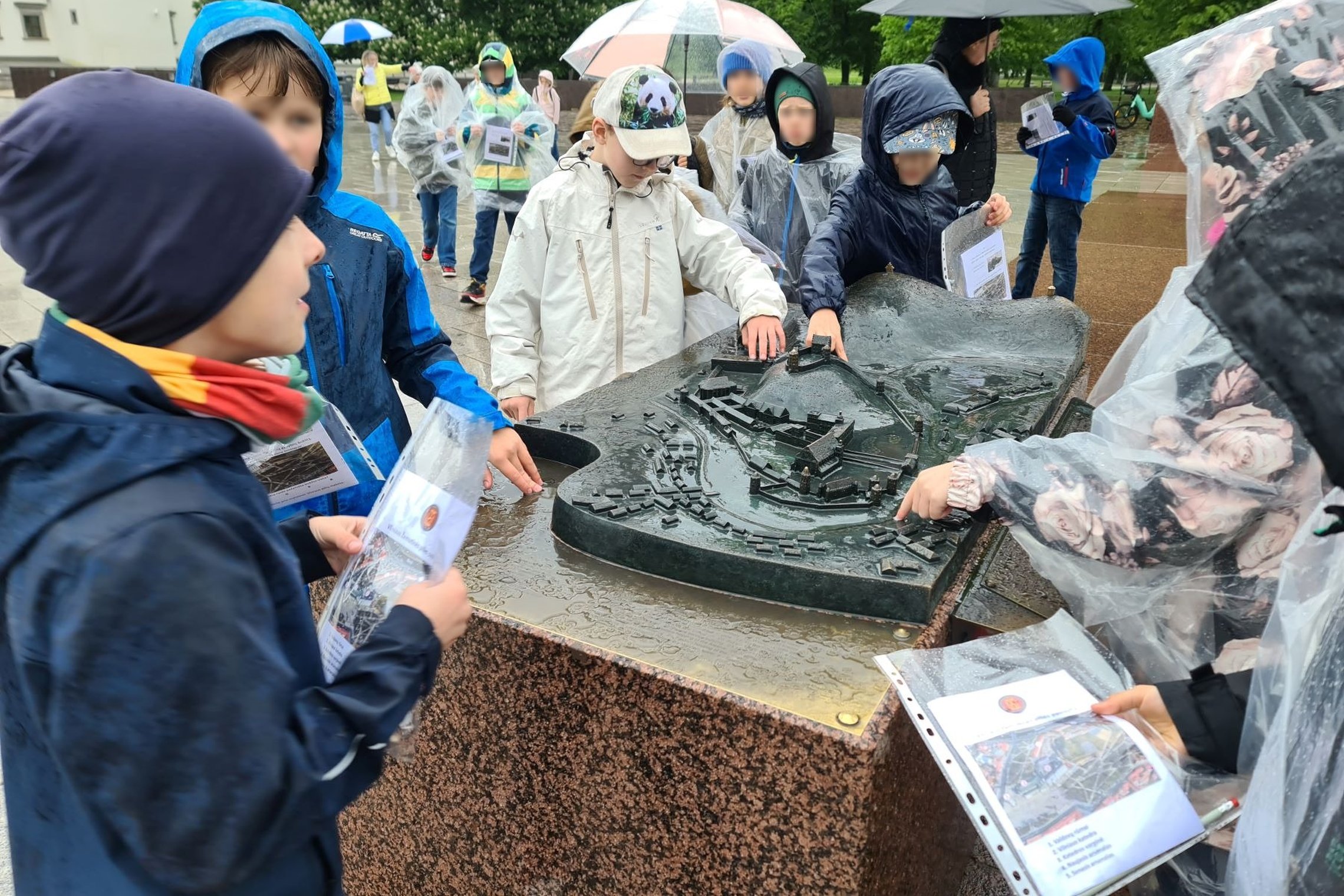 Vaikai per lietų, su lietpalčiais prie metalinio Vilniaus pilių maketo Katedros aikštėje.