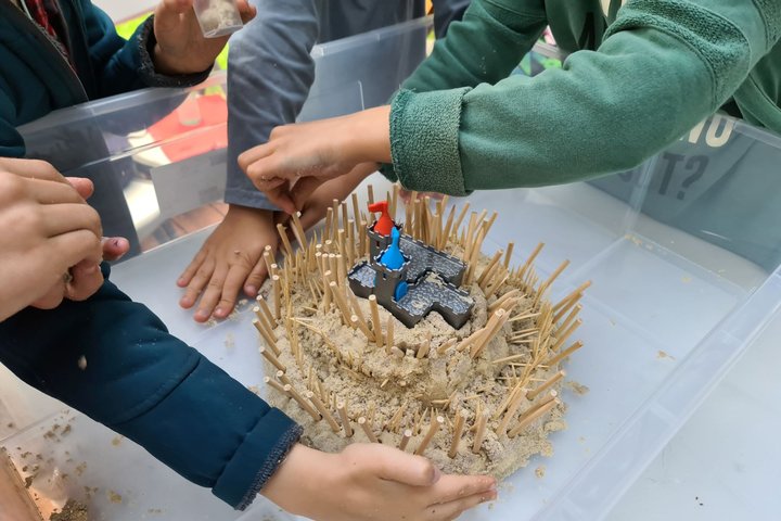 Vaikučių rankos stato smėlio pilį plastikinėje dėžėje.