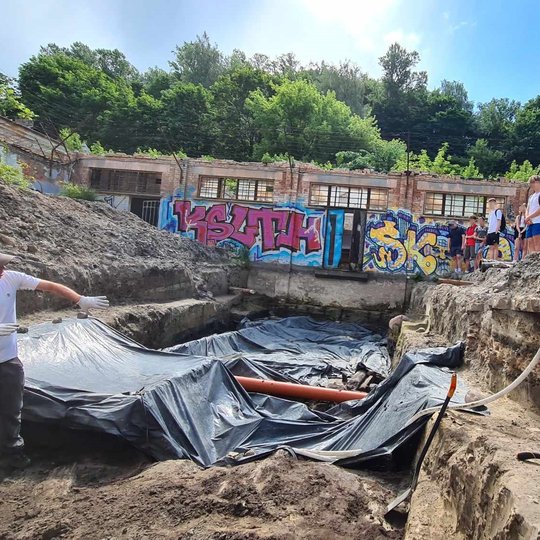 Mokiniams, stovintiems ant perkasos krašto, archeologas duobėje pasakoja ir rodo savo atradimus.