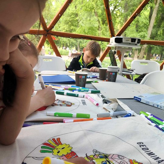 Vaikai prie stalo spalvina medžiaginius krepšius, ant kurių yra valdovų portretai.