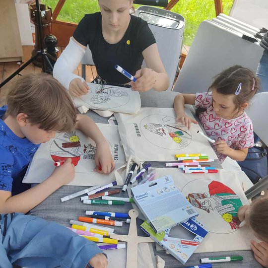 Vaikai prie stalo spalvina medžiaginius krepšius, ant kurių yra valdovų portretai.