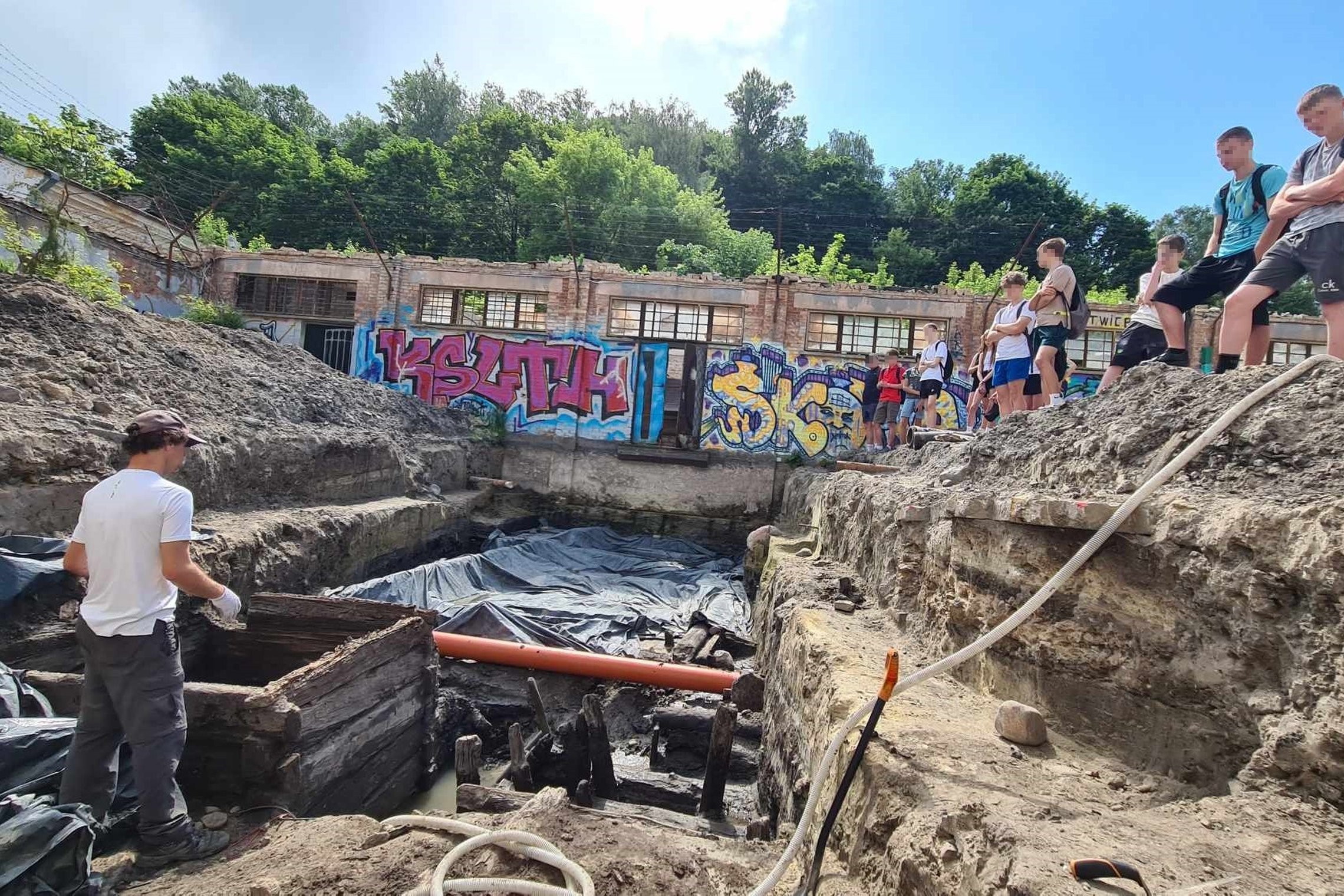 Mokiniai stovi prie perkasos krašto, o perkasoje stovi archeologas.