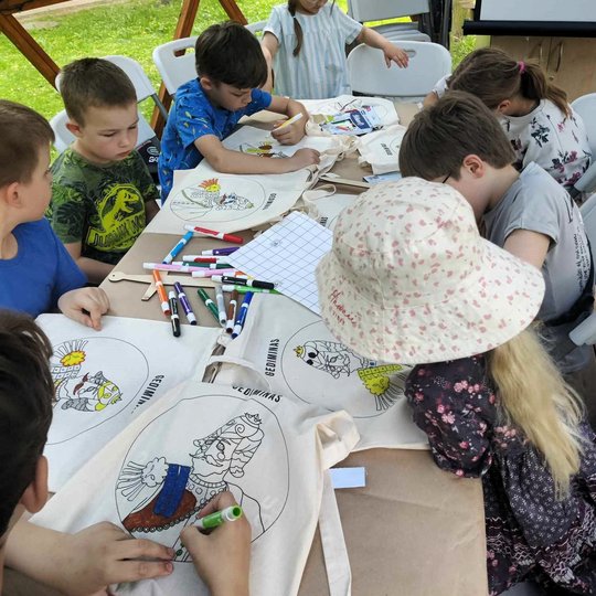 Vaikai prie stalo spalvina medžiaginius krepšius, ant kurių yra valdovų portretai.