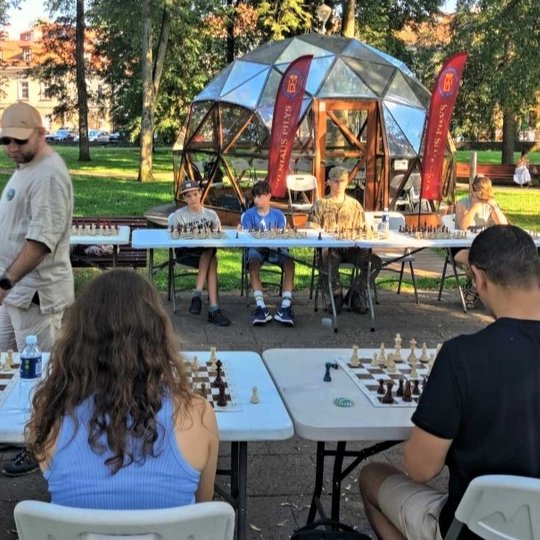 Du šachmatų stalai Sereikiškių parke, prie kurių sėdi žaidėjai. Fone matosi Vilniaus pilių kupolas.
