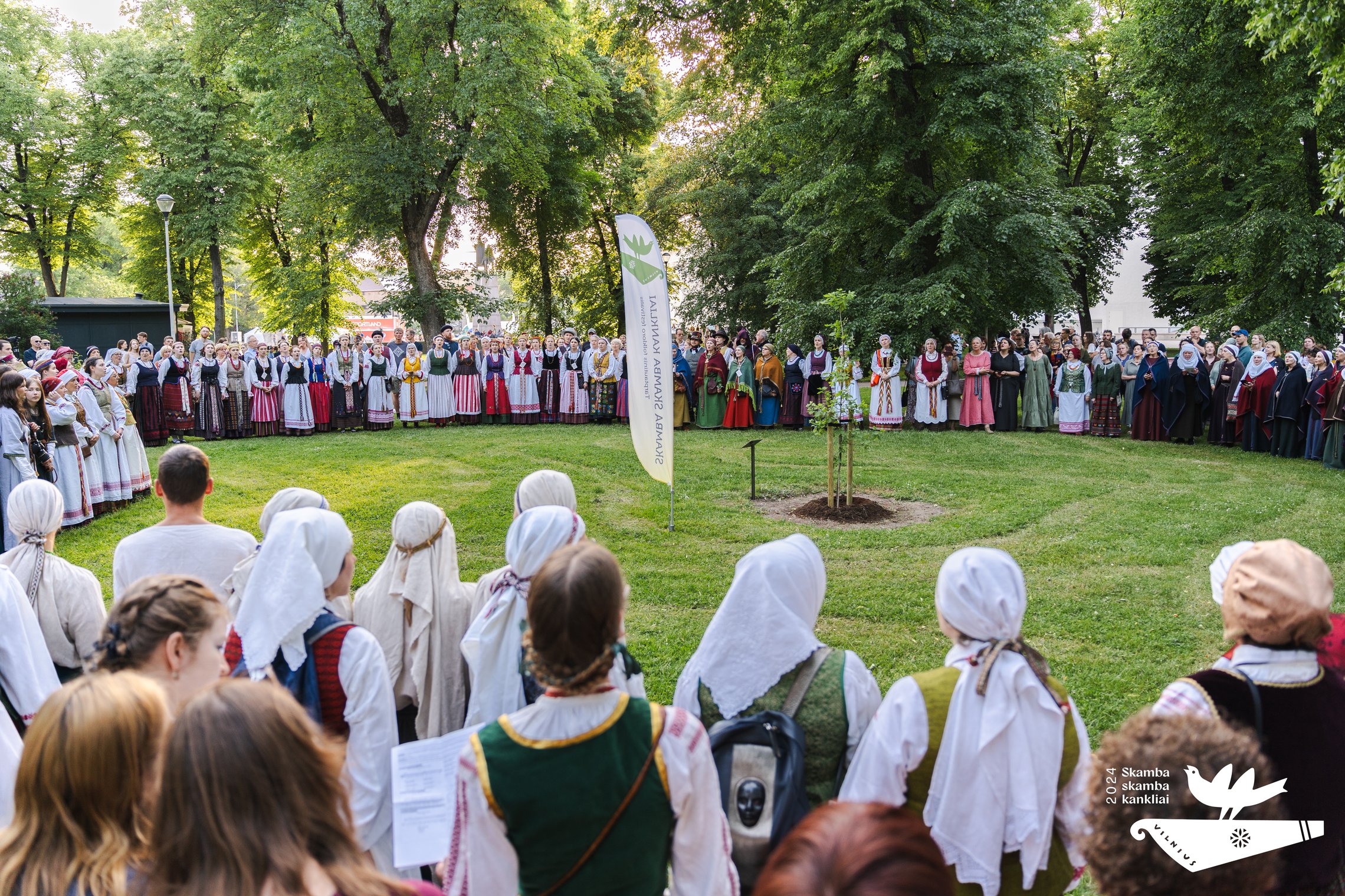 Milžiniška minia tautiniais rūbais pasipuošusių žmonių stovi ratu aplink pasodintą ąžuolą Sereikiškių parke.