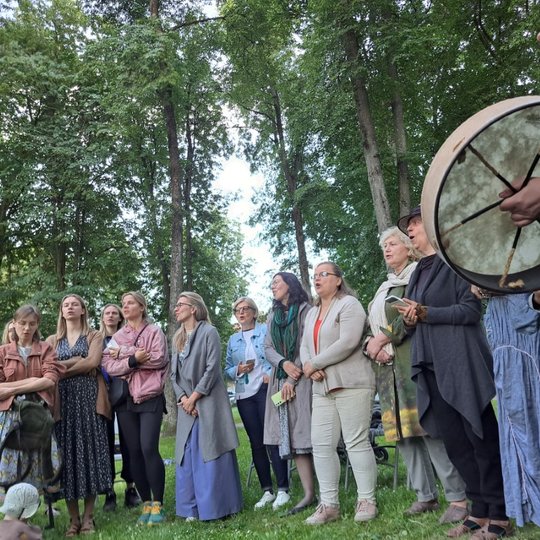 Moterys Sereikiškių parke, sustoję ratu, dainuoja sutartines. Viena moteris muša būgną.
