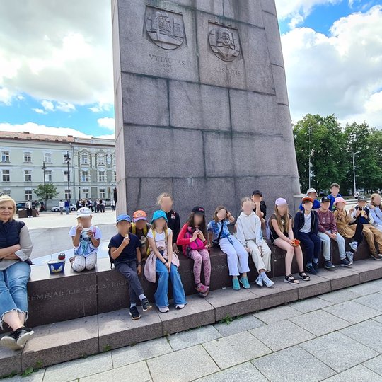 Mokiniai sėdi prie Gedimino paminklo Katedros aikštėje. Kai kurie valgo ledus.