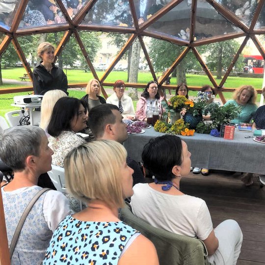 Kupole susėdę žmonės klausosi paskaitos, stovi paskaitos lektorė. Ant padengto stalo pamerktos vaistažolės.