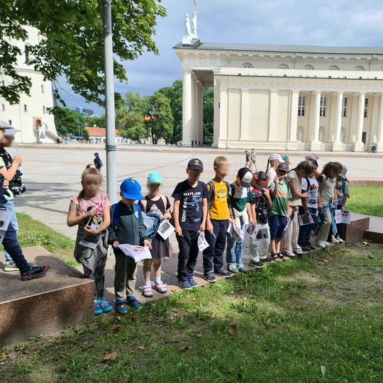 Vaikai išsirikiavę į eilę ant gynybinę sieną žyminčios plokštės Katedros aikštėje.