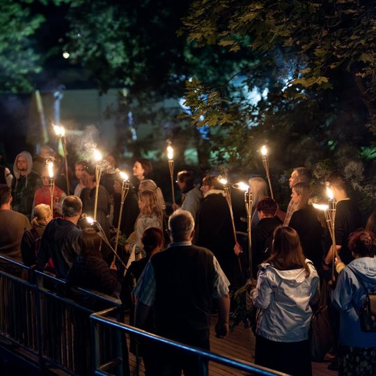 Grupė ekskursijos dalyvių su deglais rankose stovi ant tilto per Vilnios upę.