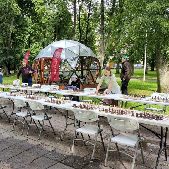 Sustatytos dvi eilės stalų su kėdėmis, ant kurių yra šachmatų lentos ir figūros, prie Vilniaus pilių kupolo.