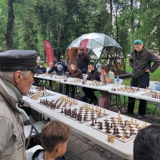 Žmonės prie pailgo stalo parke žaidžia šachmatais. Kai kurie net atsistoję.