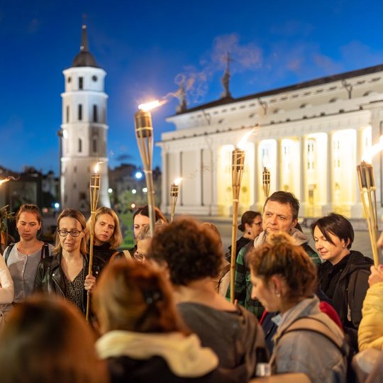 Grupė ekskursijos dalyvių su deglais rankose stovi Katedros aikštėje.