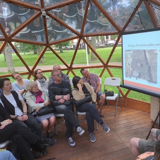 Pilnas kupolas susėdusių žmonių klausosi paskaitos. Projektoriuje matomi radiniai iš Vilniaus pilių.