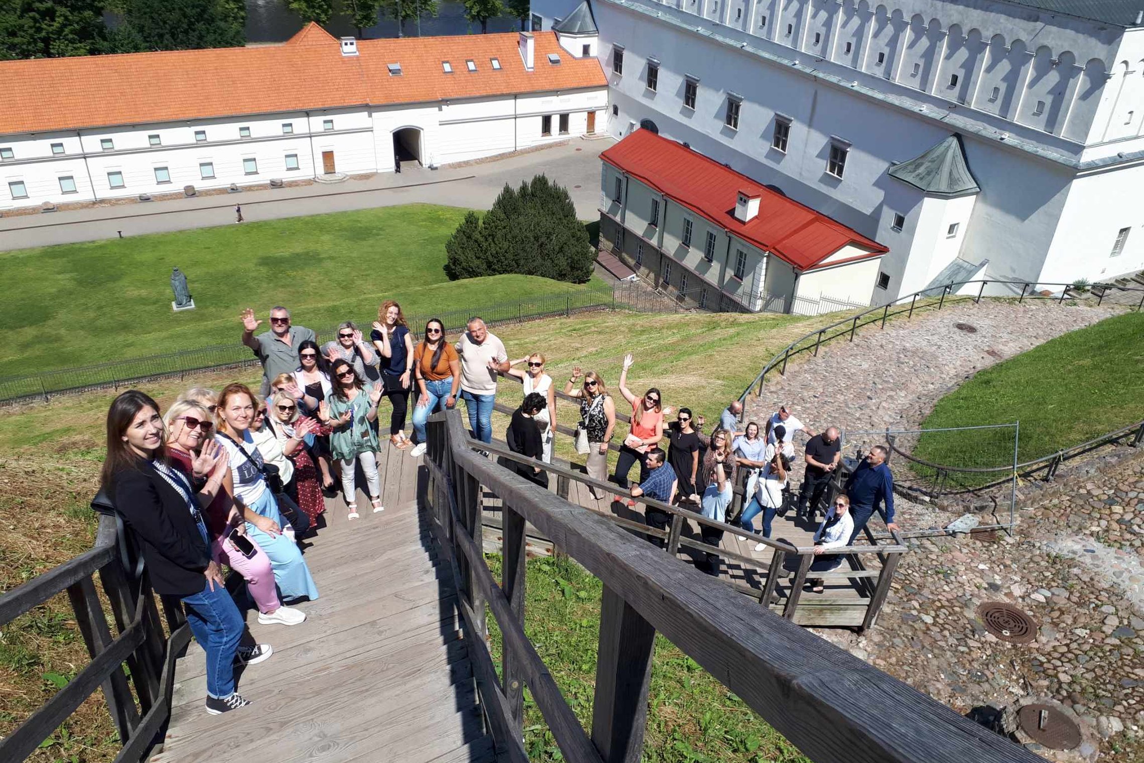 Didelė grupė ekskursijos dalyvių mojuoja kamerai nuo Gedimino pilies kalno laiptų.