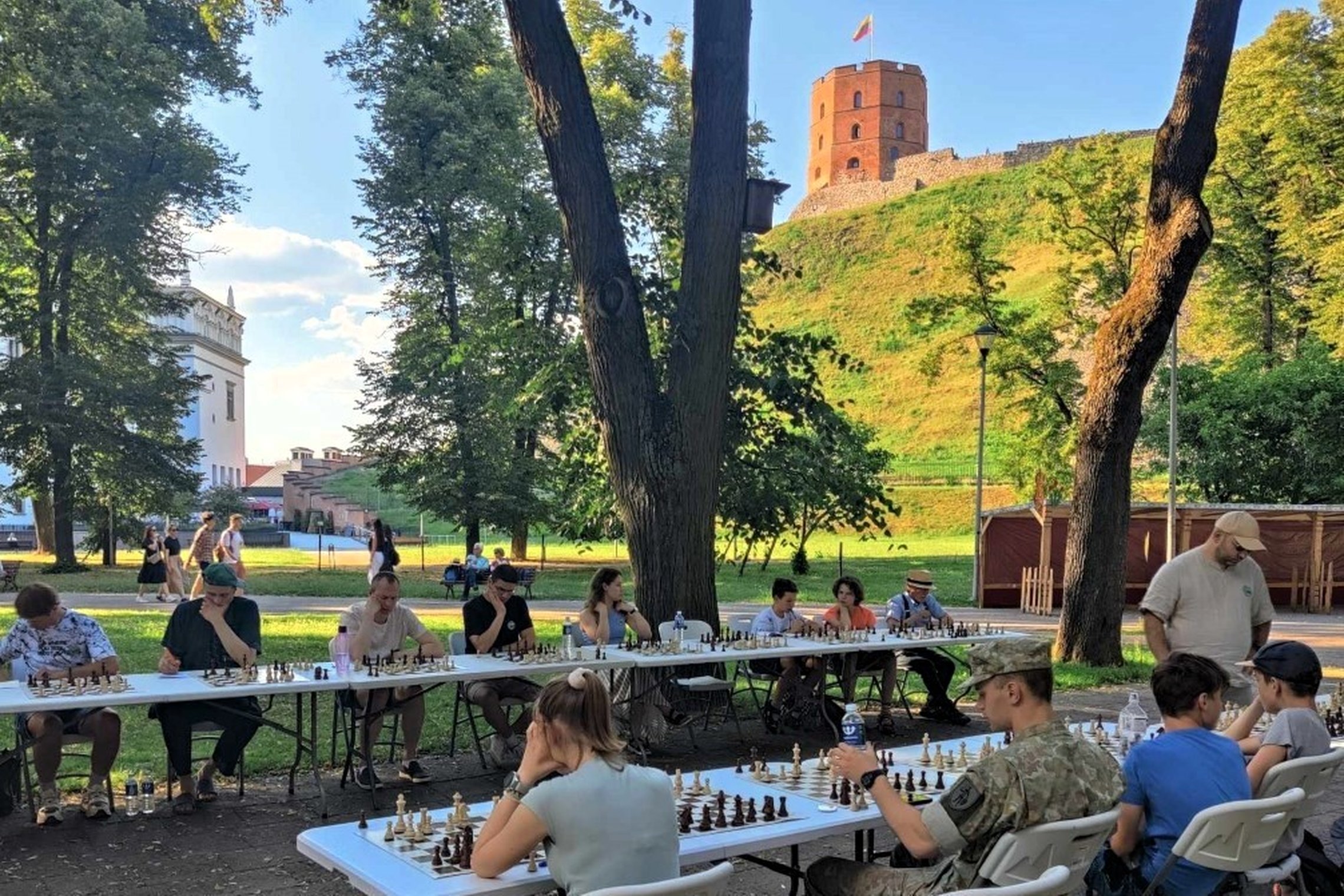 Du šachmatų stalai Sereikiškių parke, prie kurių sėdi žaidėjai. Fone matosi Gedimino pilies bokštas.