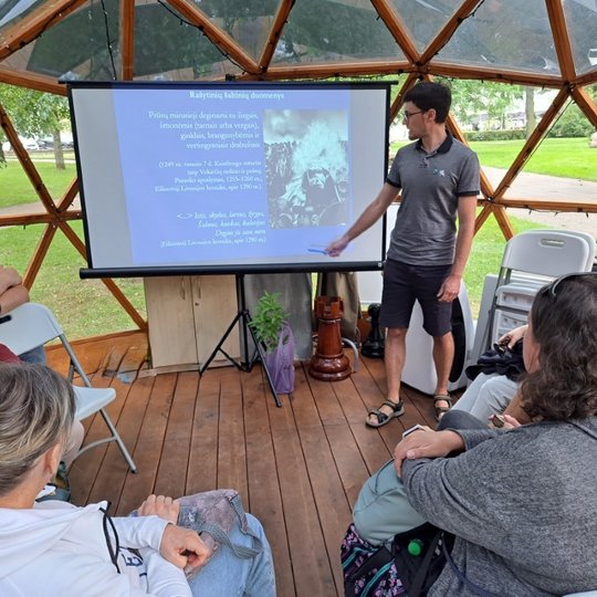 Archeologas rodo rašikliu į vaizdą projektoriuje. Aplink jį sėdi žmonės ir klausosi pasakojimo.
