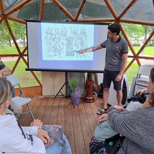 Archeologas rodo rašikliu į vaizdą projektoriuje. Aplink jį sėdi žmonės ir klausosi pasakojimo.