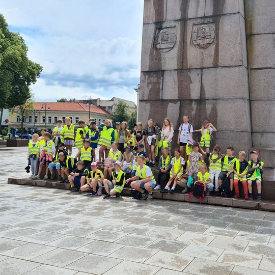 Mokinių grupė stovi Gedimino paminklo pavėsyje.