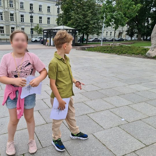 Du vaikai stovi Katedros aikštėje su užduočių lapais.