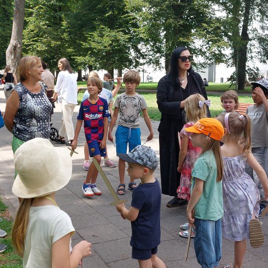 Vaikai klausosi ekskursijos Sereikiškių parke. Rankose kai kurie laiko medinius kardus.
