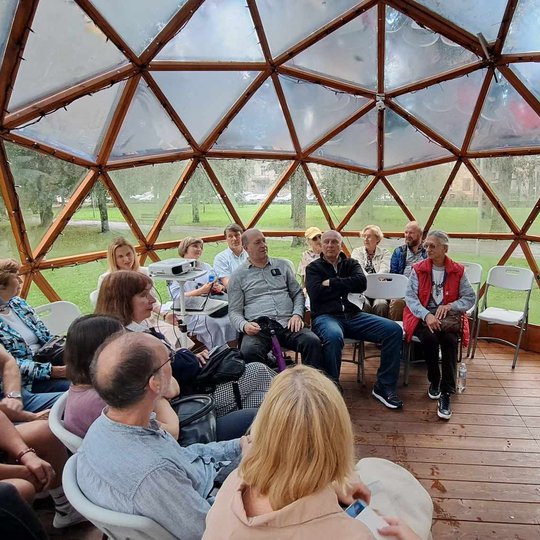 Žmonės susėdę puslankiu Vilniaus pilių kupole. Tarp jų yra ir  lektorė. Visi stebi prezentaciją projektoriaus ekrane prieš juos.