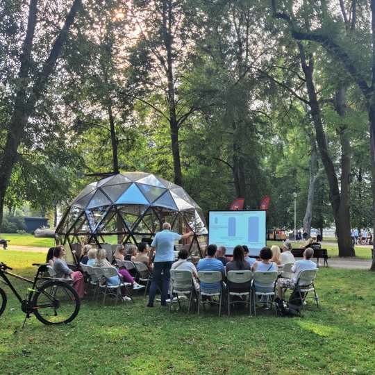 Parke, prie kupolo, klausytojai sėdi ant kėdžių, o lektorius rankose mojuoja mediniu kardu ir veda paskaitą.