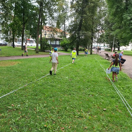 Mokiniai matuoja tariamos perkasos ilgį parko pievoje.