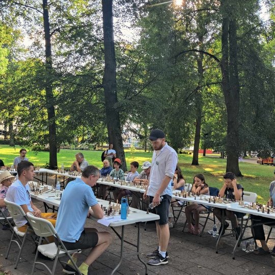 Šachmatininkai sėdi puslankiu prie stalų. Vienas žmogus stovi prie jų ir žaidžia priešininku.