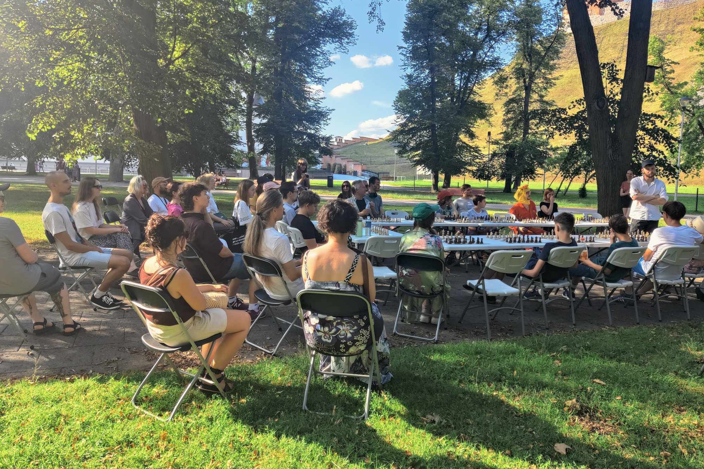 Būrys žmonių sėdi Sereikiškių parke prie šachmatų stalų.