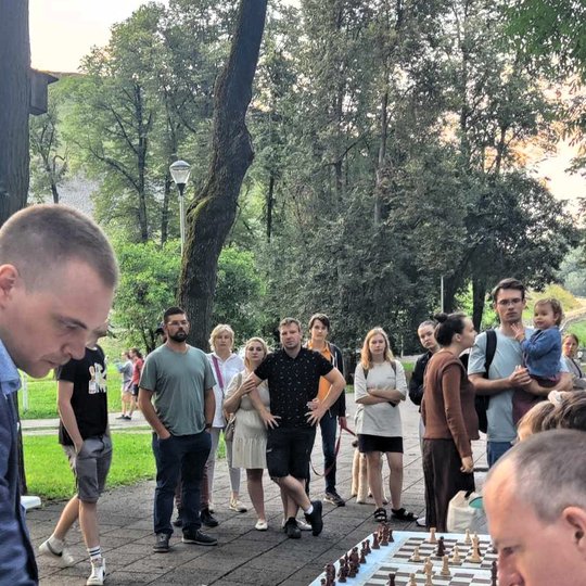 Šachmatų žaidimą stebi minia žmonių parke.