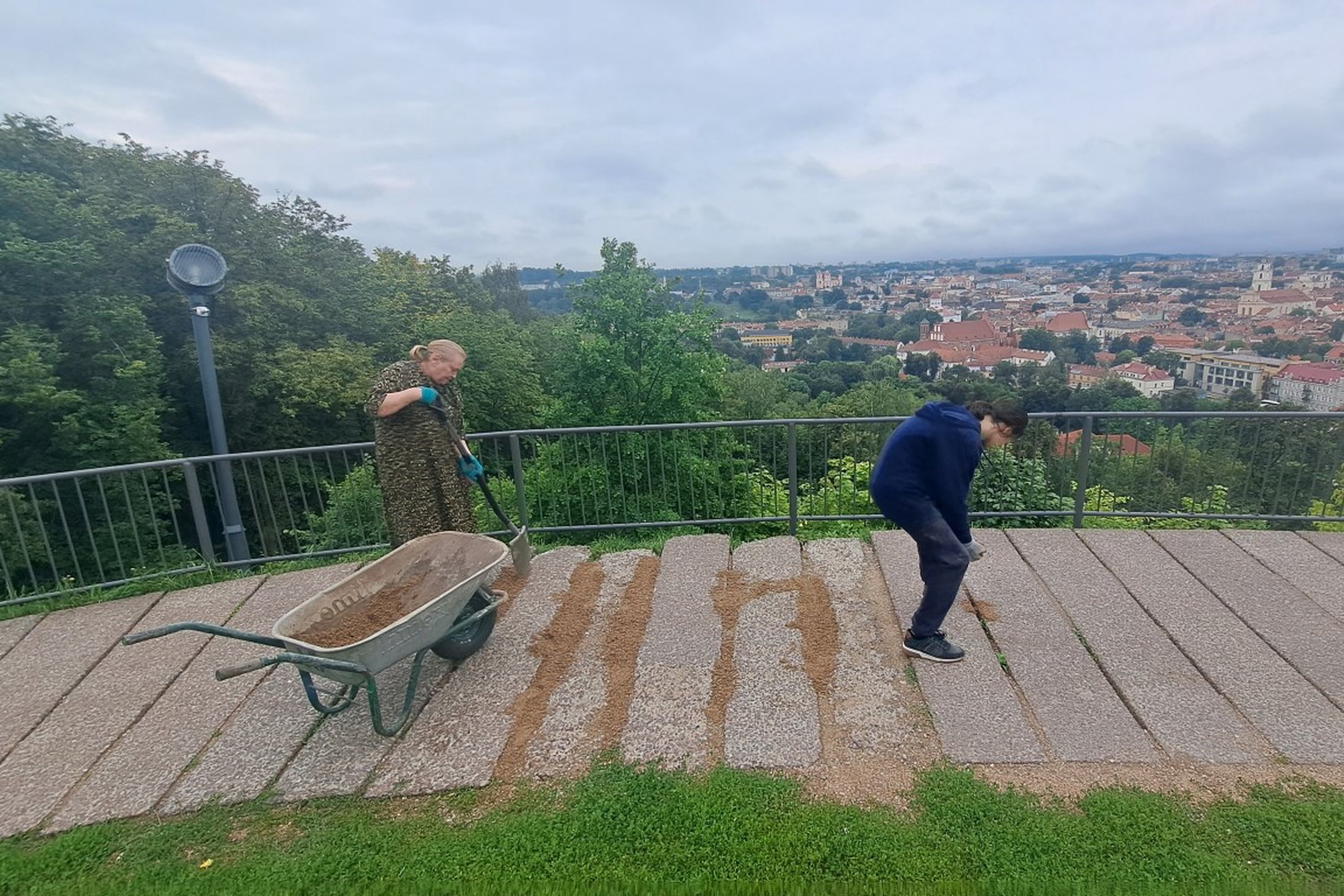 Moteris  kastuvu pila skaldą į aikštelės plyšius, o vaikinas lygina ir spaudžia skaldelę.