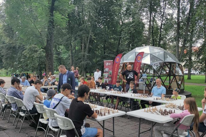 Septyni balti stalai išdėlioti Sereikiškių parke ratu. Prie stalų vyksta šachmatų žaidimų kovos.