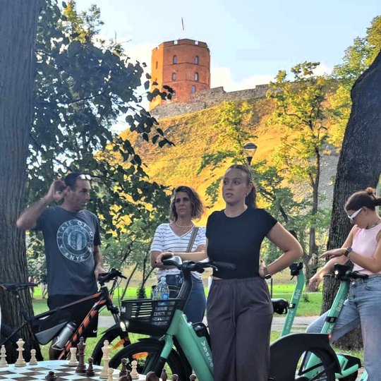 Vienas žmogus žaidžia šachmatais prie stalo. Trys žmonės stebi, stovintys prie dviračių.