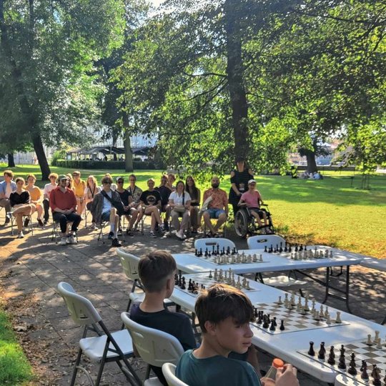 Būrys žmonių sėdi Sereikiškių parke prie šachmatų stalų. Grupė sėdinčiųjų žiūri į žaidžiančius.